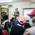 Grupo diverso participa de reunião comunitária enquanto uma pessoa apresenta informações diante de uma tela.