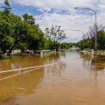 Estrada alagada com uma ponte submersa, cercada por árvores sob céu nublado.