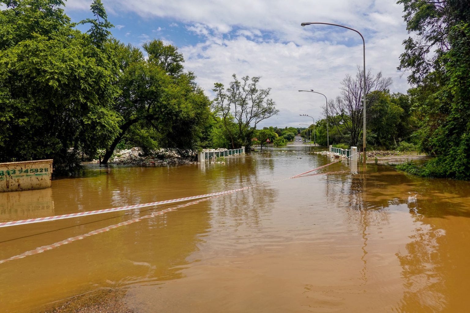 Estrada alagada com uma ponte submersa, cercada por árvores sob céu nublado.