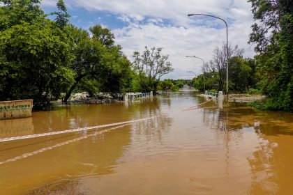 Estrada alagada com uma ponte submersa, cercada por árvores sob céu nublado.