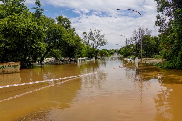 Estrada alagada com uma ponte submersa, cercada por árvores sob céu nublado.